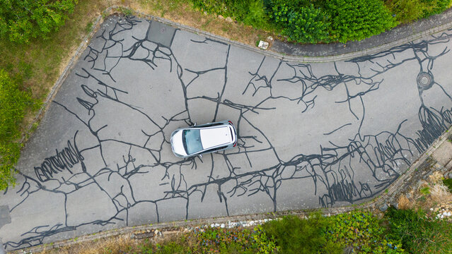 Aerial view of a silver car parked on an asphalt road with extensive crack repairs and dark tar lines in Trier-Saarburg, Rhineland-Palatinate, Germany.