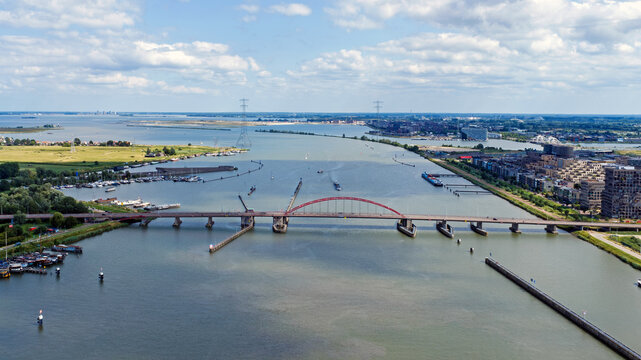 Aerial view of the Schellingwouderbrug bridge spanning the IJ river with boats, urban development, and lush green banks in Amsterdam, Noord-Holland, Netherlands.