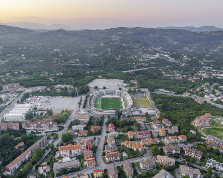 Aerial view of Stadio Partenio-Adriano Lombardi surrounded by residential buildings and lush green hills during golden hour in Avellino, Campania, Italy.