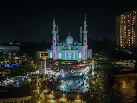 Aerial view of Jami' At-Thohir Mosque illuminated at night with colorful lights, surrounded by a bustling plaza with food stalls and visitors in Kecamatan Tapos, Jawa Barat, Indonesia.