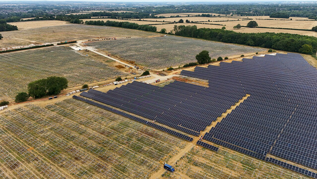 Aerial view of solar farm construction featuring rows of photovoltaic panels installed on agricultural land with surrounding fields and trees in Great Wymondley, England, United Kingdom.