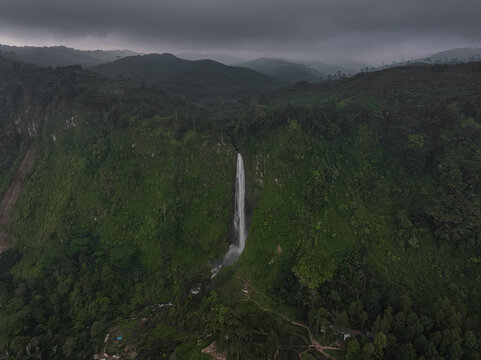 Aerial view of Citambur Waterfall cascading down a lush green cliff surrounded by dense tropical forest under a cloudy sky in Pasirkuda, West Java, Indonesia.