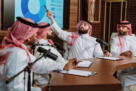 Saudi men in thobe and ghutra sit at a table with microphones