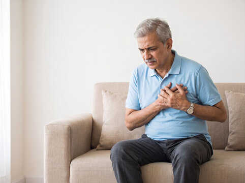 Indian senior man experiencing sudden chest pain while seated on a sofa with copy space for heart attack awareness cardiology healthcare emergency and insurance advertising