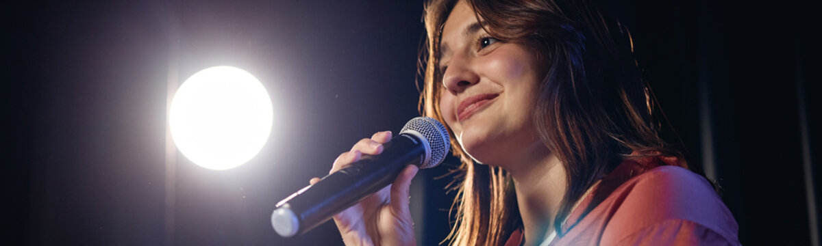 Caucasian girl holding microphone on stage during student theater performance. Image represents public speaking and arts education, ideal for school drama program promotion
