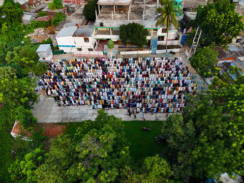 Aerial view of a large crowd of men gathered for outdoor prayer in an open courtyard near a mosque with a green dome in Kushtia, Khulna Division, Bangladesh.