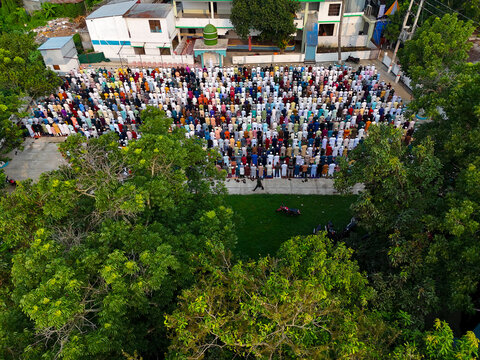 Aerial view of a large congregation of men in traditional clothing performing prayers in an outdoor courtyard surrounded by lush green trees in Kushtia, Khulna Division, Bangladesh.