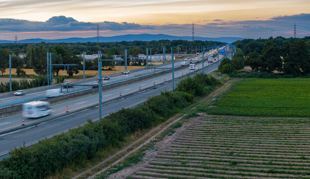 Aerial view of the eHighway test track for trucks with overhead power lines along the A5 motorway, surrounded by fields and trees under a sunset sky in Weiterstadt, Hessen, Germany.