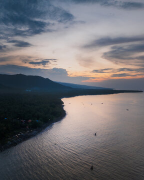 Aerial view of Amed coastline at sunset with calm waters, dark volcanic sand beaches, lush tropical vegetation, and small traditional fishing boats in Abang, Bali, Indonesia.