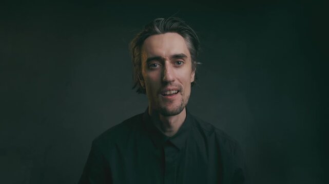 smile and nod approvingly Middle-aged man with shoulder-length hair and beard in a dark collared shirt, friendly expression in medium close-up against a dark background