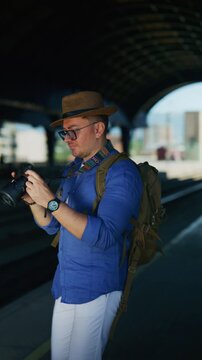 A male tourist photographer wearing a hat and glasses captures images with his digital camera. He thoughtfully reviews photos while waiting at an urban train platform.