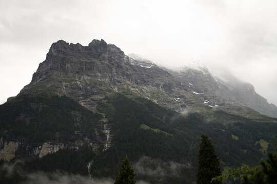 View of the Eiger mountain partially covered by clouds with pine forests and rocky slopes in Grindelwald, Canton of Bern, Switzerland.