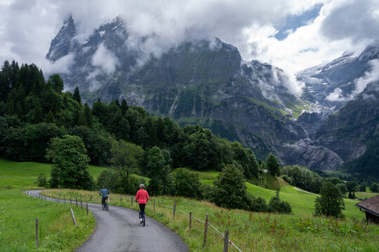 View of two cyclists riding on a path through the green meadows of Grindelwald with the dramatic Eiger mountain range in the background, Grindelwald, Canton of Bern, Switzerland.