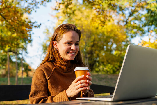 Smiling adult student enjoying online education class on laptop with takeaway coffee in autumn park