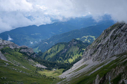 View of Mount Pilatus with rocky slopes, lush green valleys, and a paraglider soaring through the cloudy sky in Alpnach, Obwalden, Switzerland.
