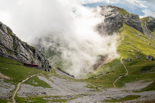 View of the Mount Pilatus cogwheel railway train traversing the rocky slopes amidst clouds and green alpine meadows in Alpnach, Obwalden, Switzerland.