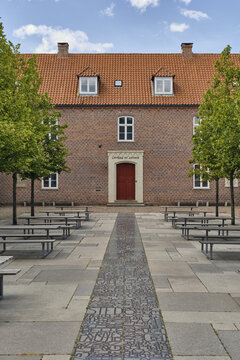 View of the historic brick building in Ribe with a red door, featuring a paved courtyard with benches and a path inscribed with text in Ribe, Syddanmark, Denmark.