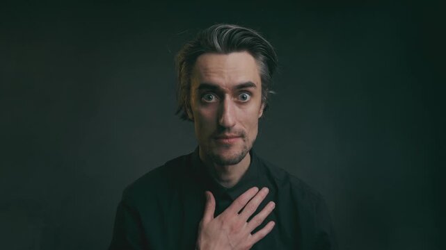 oops that's me &ndash; Close-up portrait of a young adult man in a black shirt with hand on chest, expressing contemplation against a dark neutral background