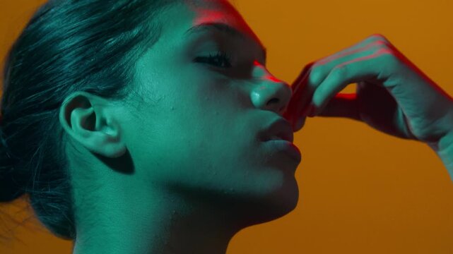 Portrait close-up of young model lit with red and cyan light touches her face with dorsal of her hand sitting in profile an orange-lit background | Fragrance product commercial
