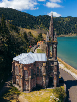 Aerial view of Ermida de Nossa Senhora das Vitorias, a neo-Gothic chapel with a tall spire on the shore of Furnas Lake, surrounded by lush hills in Furnas, Acores, Portugal.