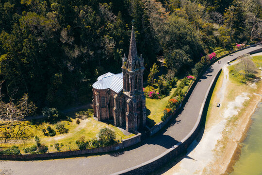 Aerial view of the neo-Gothic Ermida de Nossa Senhora das Vitorias chapel on the shore of Furnas Lake, surrounded by lush forest and vibrant flowers in Furnas, Acores, Portugal.