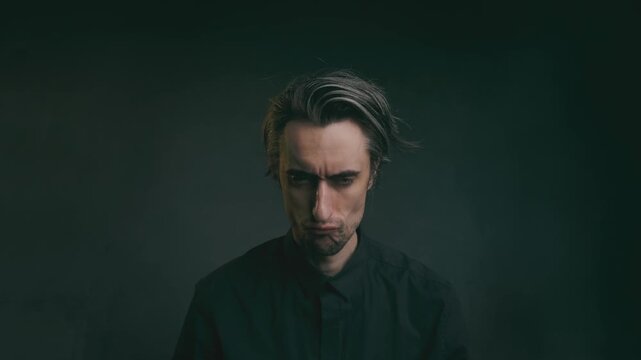 serious comical frowning sulking offended Moody studio portrait of a solitary young man in a dark collared shirt, head lowered, with tousled hair against a muted dark gradient background