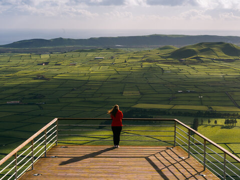 View of a person standing on a wooden observation deck overlooking the vast green patchwork fields of Terceira Island in the Azores, Portugal.