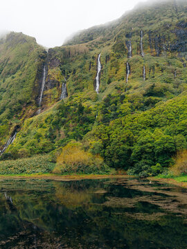 View of the lush green cliffs of Flores Island featuring multiple cascading waterfalls flowing into a calm pond in the Azores, Portugal.