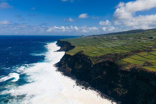 Aerial view of Terceira Island featuring dramatic volcanic cliffs, crashing waves, and lush green agricultural fields under a clear blue sky in Azores, Portugal.