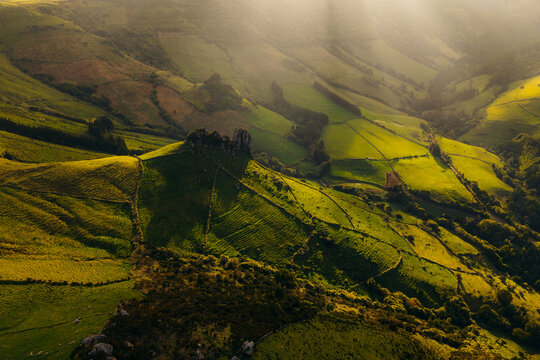 Aerial view of the lush green rolling hills and rocky outcrops of Flores Island, defined by traditional stone walls under the warm glow of the golden hour in Azores, Portugal.