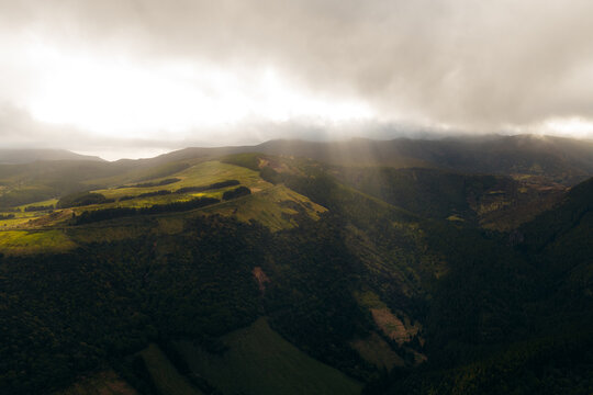 Aerial view of Flores Island in the Azores, Portugal, featuring lush green rolling hills, deep valleys, and dramatic sunbeams breaking through heavy clouds over the rugged landscape.