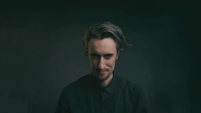 The cunning one puffed up, smiling wickedly, a madman showing serious to sinister expressions in a dark button-up shirt against a dim gray-black background with chiaroscuro lighting.