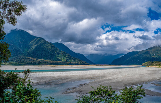 Haast River, Mount Aspiring National Park, breites Flussbett mit tuerkisfarbenem Wasser, Wolken haengen tief ueber bewaldeten Bergen, Wanderweg im Vordergrund, Suedinsel, Neuseeland