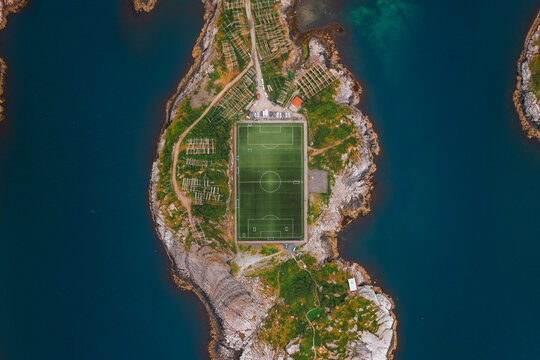 Aerial view of Henningsvaer Football Stadium on a rocky island surrounded by deep blue water, featuring fish drying racks and rugged coastal terrain in Henningsvaer, Nordland, Norway.