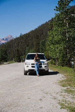 View of a woman leaning against a white SUV on a gravel road with mountains and pine trees in the background in Canmore, Alberta, Canada.