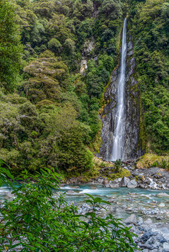 Thunder Creek Falls, Mount Aspiring National Park, Haast Pass, Wasserfall stuerzt durch dichten Regenwald, tuerkisfarbener Fluss mit Felsbrocken im Vordergrund, Suedinsel, Neuseeland