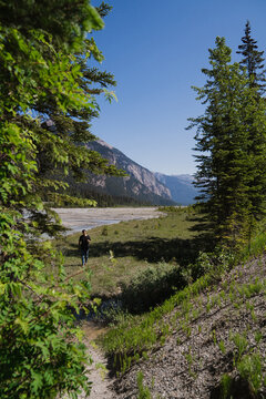 View of a person walking along a grassy path near a river with mountains in the background under a clear blue sky in Canmore, Alberta, Canada.