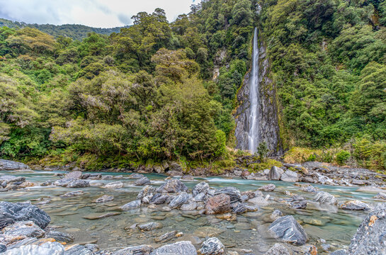 Thunder Creek Falls, Mount Aspiring National Park, Haast Pass, Wasserfall stuerzt durch dichten Regenwald, tuerkisfarbener Fluss mit Felsbrocken im Vordergrund, Suedinsel, Neuseeland