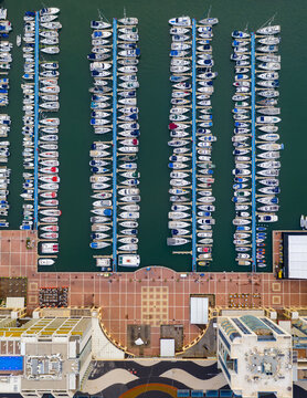 Aerial view of Marina Herzliya with rows of yachts and sailboats docked at the piers, next to a paved promenade and modern waterfront buildings in Herzliya, Tel Aviv District, Israel.