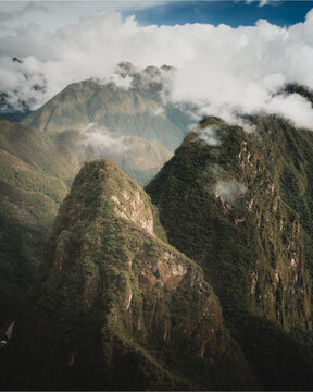 Aerial view of lush, cloud-covered Andes mountain peaks, featuring steep, vegetation-covered slopes and dramatic rocky ridges under a soft, overcast sky in Aguas Calientes, Cusco, Peru.
