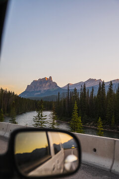 View of the majestic mountain peaks reflecting in a car side mirror during golden hour with a river and pine forest in Lake Louise, Alberta, Canada.