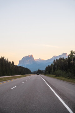 View of a scenic highway leading towards the majestic mountains of the Canadian Rockies under a soft sunset sky in Lake Louise, Alberta, Canada.
