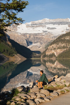 View of a man sitting on the rocky shore of Lake Louise, enjoying the reflection of the snow-capped mountains and pine forests in the calm water, Lake Louise, Alberta, Canada.
