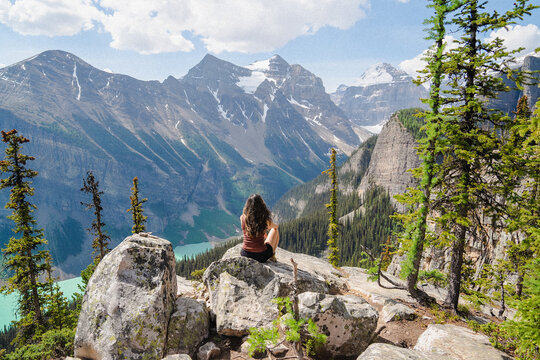 View of a woman sitting on a rocky outcrop overlooking the turquoise waters of Lake Louise surrounded by majestic snow-capped mountains and evergreen pine forests in Lake Louise, Alberta, Canada.