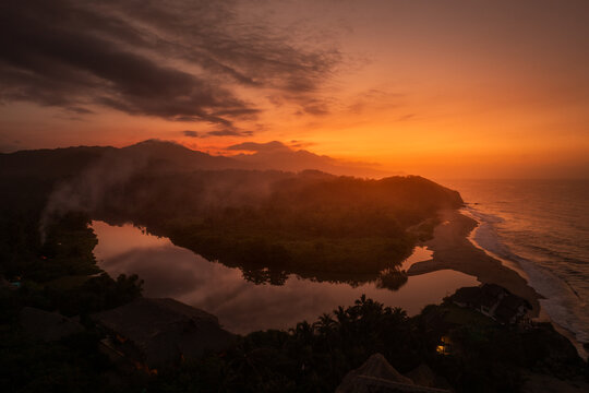 Aerial view of Parque Nacional Natural Tayrona at sunset, featuring a calm river meeting the Caribbean Sea, lush tropical forest, and dramatic orange sky in Magdalena, Colombia.