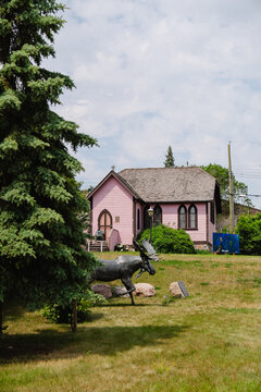 View of a pink church with a wooden shingle roof, set in a grassy park with a bronze moose statue and a large evergreen tree in Brandon, Manitoba, Canada.