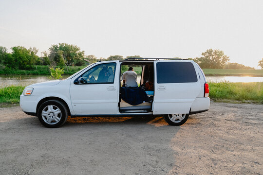 View of a white minivan parked by a river with a person sitting inside during the golden hour in Brandon, Manitoba, Canada.