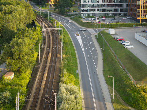 Aerial view of railway tracks running parallel to a road with modern buildings, parked cars, and lush greenery in Krakow, Lesser Poland Voivode, Poland.