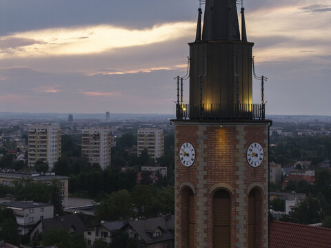 Aerial view of the Church of Our Lady of Good Counsel in Prokocim with its brick clock tower illuminated against a sunset sky, overlooking residential Krakow, Malopolskie, Poland.