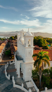 Aerial view of Santa Lucia Church with its white facade and bell towers, surrounded by red-tiled rooftops, lush palm trees, and distant mountains in Suchitoto, Cuscatlan Department, El Salvador.